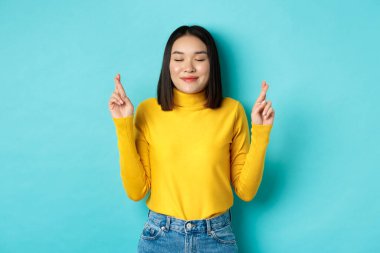 Umut dolu Asyalı kızın portresi gerçekleşiyor, şans diliyor, gülümsüyor, Tanrı 'ya yalvarıyor, mavi arka planda duruyor.