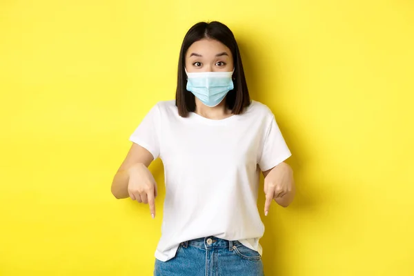 Covid-19, conceito de pandemia e distanciamento social. Incrível mulher asiática em máscara médica, mostrando propaganda, apontando para a direita e sorrindo, fundo amarelo — Fotografia de Stock