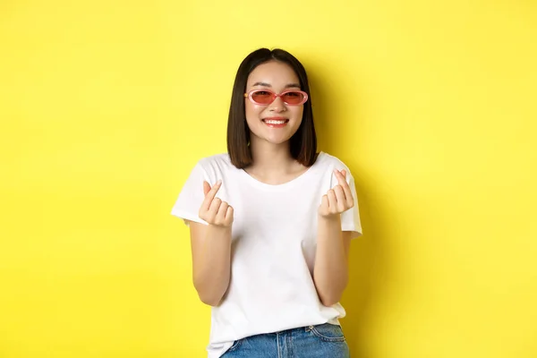 Concepto de moda y estilo de vida. Atractiva mujer asiática en gafas de sol con estilo, mostrando los corazones de los dedos y sonriendo feliz en la cámara, de pie sobre el fondo amarillo —  Fotos de Stock