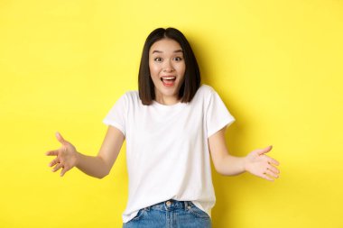 Heyecanlı genç Asyalı kadın, büyük, büyük bir objeyi tutuyormuş gibi ellerini uzattı, boyunu gösteriyor ve şaşırmış görünüyor, sarı arka planda duruyordu.