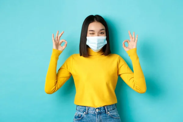 Covid-19, socialt avståndstagande och pandemikoncept. Glada asiatiska flicka i medicinsk mask visar okej gester, blinkar vid kameran, visar godkännande, står över blå bakgrund — Stockfoto