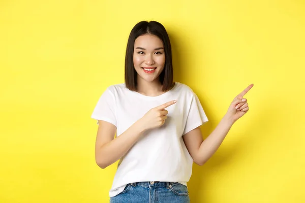 Ομορφιά και μόδα έννοια. Όμορφη ασιατική γυναίκα σε λευκό t-shirt δείχνοντας τα δάχτυλα δεξιά, δείχνουν το λογότυπο στέκεται πάνω από κίτρινο φόντο — Φωτογραφία Αρχείου