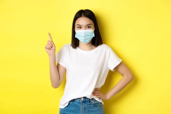 Ковед, охорона здоров'я та пандемія. Азійська жіноча модель в медичній масці і біла футболка вказівний палець у лівому верхньому лівому кутку логотип, показуючи просування, жовтий фон — стокове фото