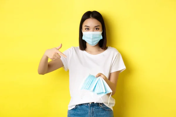 Concepção de coronavírus, quarentena e medicina. Jovem mulher asiática apontando dedo para máscaras médicas para ir ao ar livre, de pé sobre fundo amarelo — Fotografia de Stock