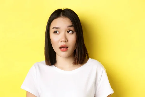 Close up de bela mulher asiática com pele perfeita limpa, levantando sobrancelha e olhando para a esquerda com rosto intrigado, de pé sobre fundo amarelo — Fotografia de Stock