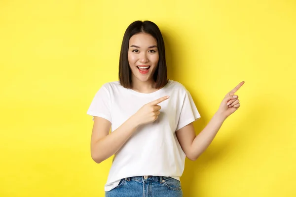 Güzellik ve moda konsepti. Beyaz tişörtlü güzel Asyalı kadın parmaklarını sağa göstererek sarı arka planda duran logoyu gösteriyor. — Stok fotoğraf