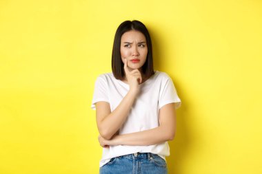 Güzellik ve moda konsepti. Düşünceli Asyalı kadın bir şeyi düşünürken düşünceli görünüyor, sarı arka planda duruyor.