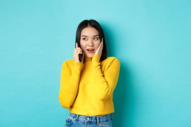 Cep telefonuyla konuşan bir kadın ilginç görünüyor. Arama sırasında ilginç bir teklif alıyor. Mavi arka planda duruyor.