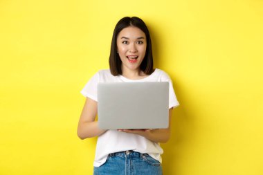 Neşeli Asyalı kadın dizüstü bilgisayarda çalışıyor ve gülümsüyor, beyaz tişört ve kot pantolonla sarı arka planda duruyor.