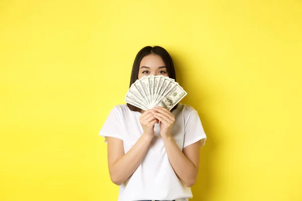 Милая азиатка, прячущая лицо за деньгами, подглядывающая в камеру довольная, зарабатывающая деньги, стоящая на жёлтом фоне — стоковое фото