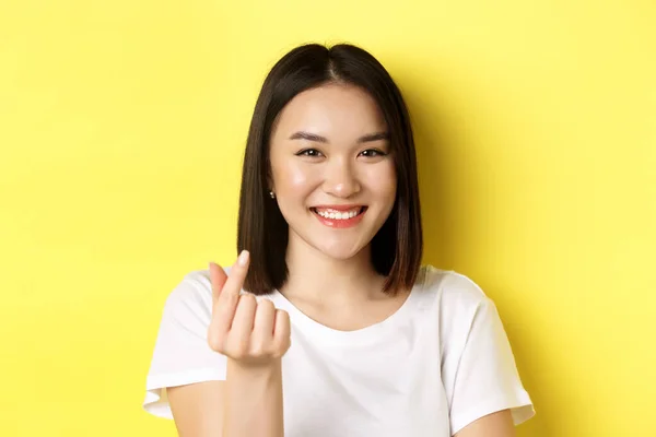 Día de San Valentín y concepto de mujeres. Primer plano de chica asiática bonita en camiseta blanca, sonriendo y mostrando el corazón del dedo, de pie sobre fondo amarillo — Foto de Stock