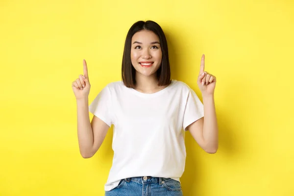 Concepto de belleza y moda. Hermosa mujer asiática en camiseta blanca apuntando con los dedos hacia arriba, de pie sobre fondo amarillo —  Fotos de Stock