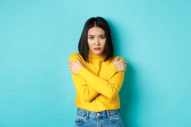 Kendini kurban gibi hisseden Asyalı genç bir kadın, elleri ile vücudu örterek kameraya endişeli bir şekilde bakıyor, mavi arka planda duruyor.