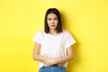 Asyalı bir kadının midesinde ağrı, karın ağrısı, acıdan çatık kaşlar, regl döneminde rahatsızlık, sarı arka planda durması.