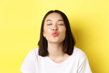 Sevgililer Günü konsepti. Tatlı Asyalı dudaklarını büzen dudaklarını ve öpmek için gözlerini kapatan, sarı arka planda romantik duran