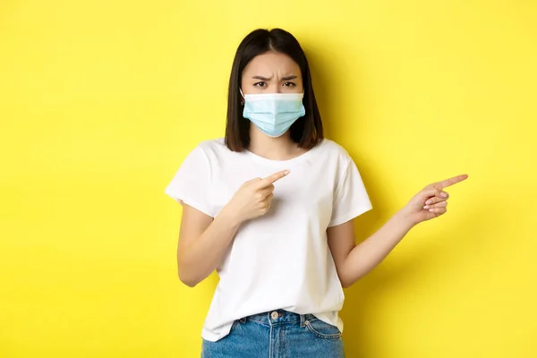 Covid-19, pandemiskt och socialt distanserande koncept. Besviken asiatisk flicka i medicinsk mask, rynkar pannan upprörd och pekar finger rätt på logotypen, står över gul bakgrund — Stockfoto