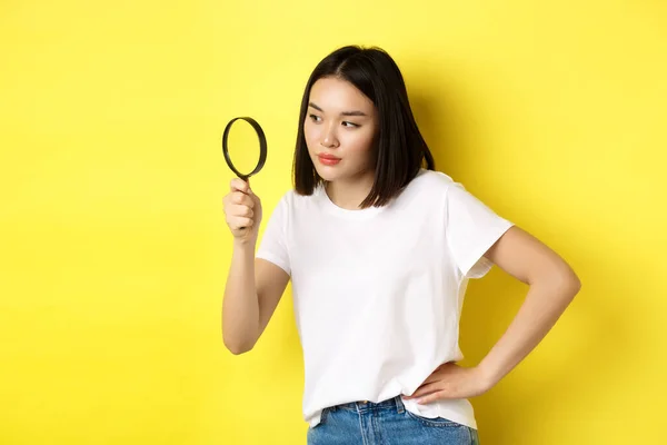 Asiatisk kvinna detektiv tittar genom förstoringsglas med spännande utseende, hittade ledtrådar, stående över gul bakgrund — Stockfoto