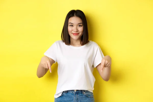 Concepto de belleza y moda. Hermosa mujer asiática en camiseta blanca apuntando con los dedos hacia abajo, demostrar el logotipo de pie sobre el fondo amarillo — Foto de Stock