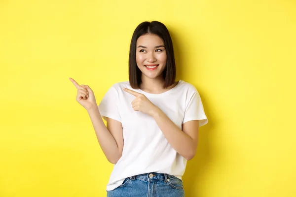 Концепція краси та моди. Красива азіатська жінка в білій футболці, спрямованій пальцями ліворуч, стоїть над жовтим тлом — стокове фото