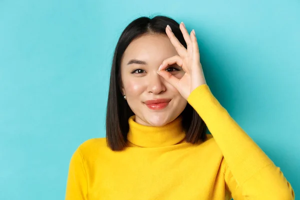 Skønhed og makeup koncept. Close up af ubekymret asiatisk pige viser OK tegn på øje og smil, ser glad på kameraet, stående over blå baggrund - Stock-foto