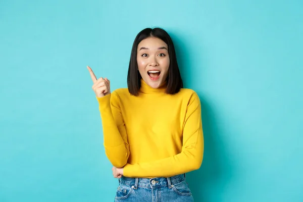 Concepto de compras. Atractiva mujer coreana sonriendo asombrada, señalando con el dedo a la izquierda, mostrando una buena pancarta, de pie sobre fondo azul — Foto de Stock