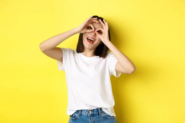 Funny asian girl looking through hand binoculars and smiling, stading over yellow background — Stock Photo, Image
