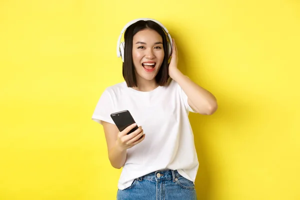 Cool chica asiática bailando y escuchando música en auriculares inalámbricos, sosteniendo el teléfono inteligente en la mano, de pie sobre fondo amarillo — Foto de Stock