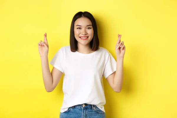 Esperançoso asiático menina fazendo desejo, cruz dedos para boa sorte e orando com os olhos fechados, de pé sobre amarelo fundo — Fotografia de Stock