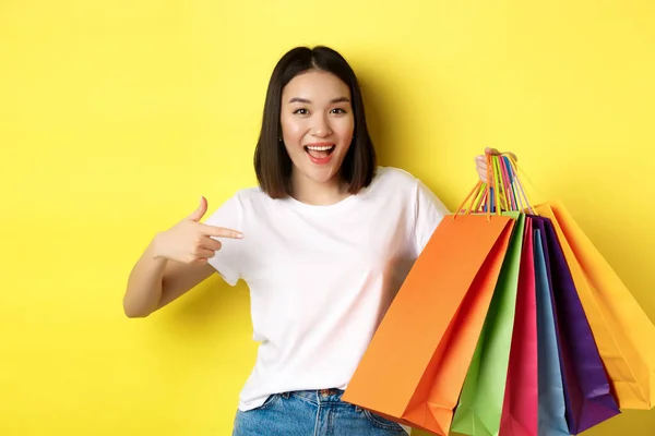 Veselá asijská žena jde na nákupy, ukazuje prstem na papírové tašky a usmívá se, nákupčí se baví při slevách, žluté pozadí — Stock fotografie