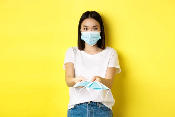 Coronavirus, καραντίνα και ιατρική έννοια. Χαριτωμένο κορίτσι της Ασίας δίνοντας ιατρικές μάσκες και κοιτάζοντας κάμερα, κίτρινο φόντο — Φωτογραφία Αρχείου