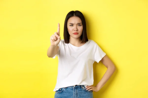 Confident and serious woman tell no, showing extended finger to stop and prohibit something bad, frowning and looking at camera self-assured, standing over yellow background — Stock Photo, Image