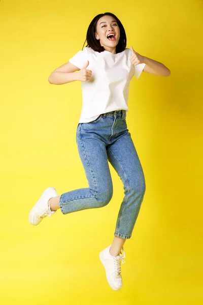 Full size of happy young asian woman jumping from joy, mostrando polegares para cima na aprovação, posando sobre fundo amarelo em jeans e casual white t-shirt — Fotografia de Stock