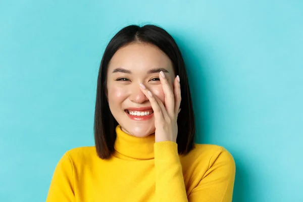 Concept beauté et maquillage. Gros plan de belle femme asiatique rougissant et riant, touchant une peau saine éclatante, souriant heureux, debout sur fond bleu — Photo