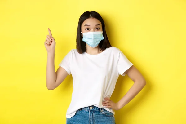 Covid, conceito de saúde e pandemia. Modelo feminino asiático em máscara médica e branco t-shirt apontando dedo no canto superior esquerdo logotipo, mostrando promoção, fundo amarelo — Fotografia de Stock