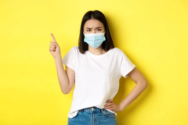 Covid, υγειονομική περίθαλψη και πανδημία έννοια. Ασιατικό γυναικείο μοντέλο με ιατρική μάσκα και λευκό t-shirt που δείχνει το δάχτυλο στο λογότυπο της πάνω αριστερής γωνίας, που δείχνει την προώθηση, κίτρινο φόντο — Φωτογραφία Αρχείου