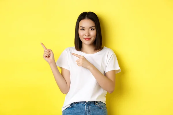 Concepto de belleza y moda. Hermosa mujer asiática en camiseta blanca señalando los dedos a la izquierda, de pie sobre fondo amarillo —  Fotos de Stock