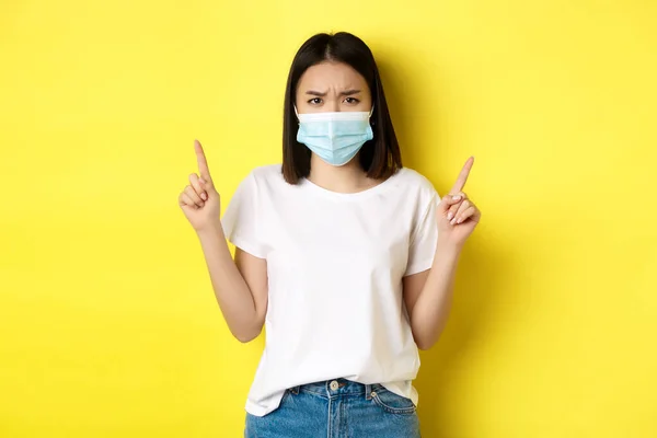 Концепція пандемії та соціального дистанціювання Covid-19. Розчарована азіатська дівчина в медичній масці, розчаровується і вказує пальці вгору за логотипом, стоячи над жовтим тлом — стокове фото