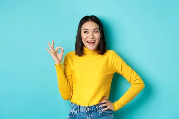 Femme asiatique impressionnée en pull jaune, regardant à gauche étonné et montrant OK signe dans l'approbation, debout sur fond bleu — Photo