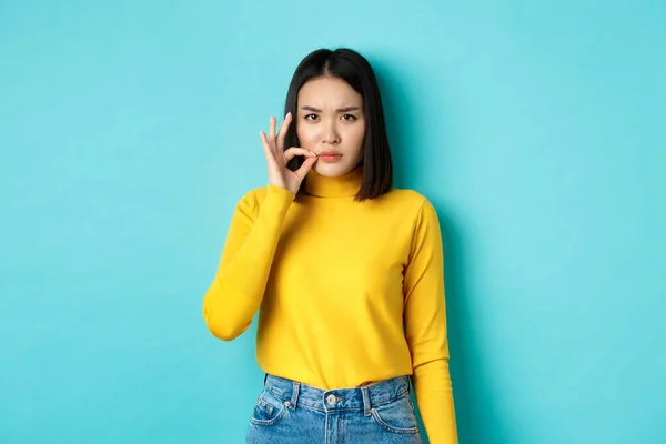 Poważny azjatycki dziewczyna pokazując usta suwak gest, obietnica trzymać zamknięty i marszczyć brwi, opowiadając duży sekret, stojąc w żółty pullover na niebieskim tle — Zdjęcie stockowe