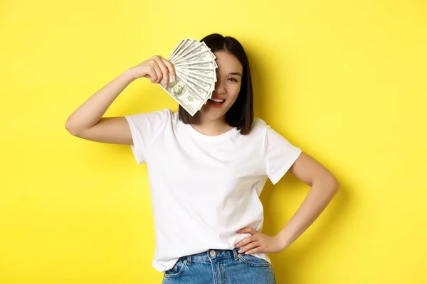 Милая азиатка, прячущая лицо за деньгами, подглядывающая в камеру довольная, зарабатывающая деньги, стоящая на жёлтом фоне — стоковое фото