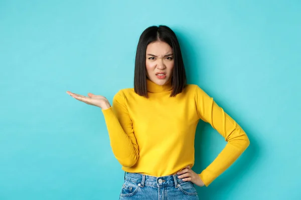 Frustrated and pissed-off asian girl raising hand up and grimacing bothered, staring at something with annoyance and disappointment, standing over blue background — Stock Fotó