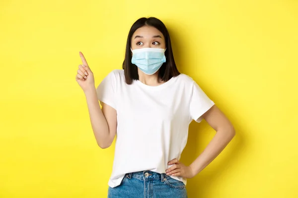 Covid, sjukvård och pandemi koncept. Asiatisk kvinna i medicinsk mask och vit t-shirt pekar finger på övre vänstra hörnet logotyp, visar befordran, gul bakgrund — Stockfoto