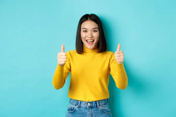 Beautiful asian woman praise good work, showing thumbs up gesture and smiling in approval, recommend product, standing satisfied over blue background