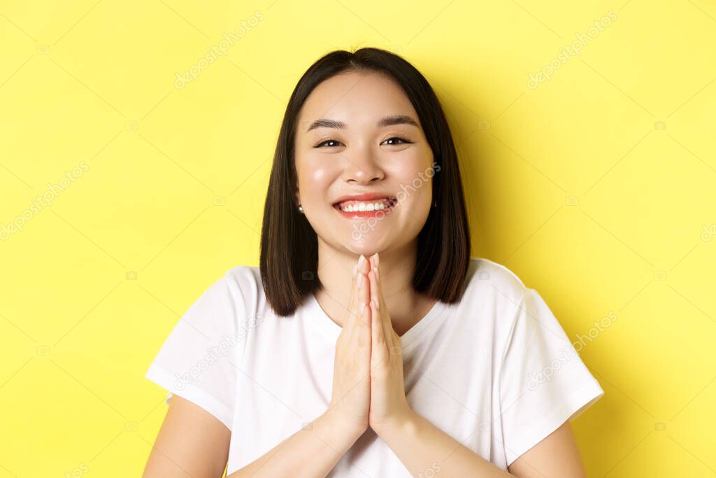 Close up of cute asian woman saying thank you and smiling, holding hands in namaste, pray gesture, standing over yellow background