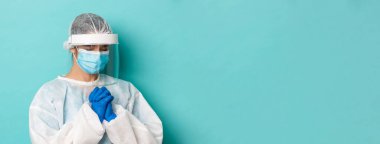 Covid-19, salgın ve karantina kavramı. Kişisel koruyucu ekipmanlarla umut dolu kadın doktorların yakın çekimleri, dua ederken, yalvarırken, yalvarırken, mavi arka planda dururken.