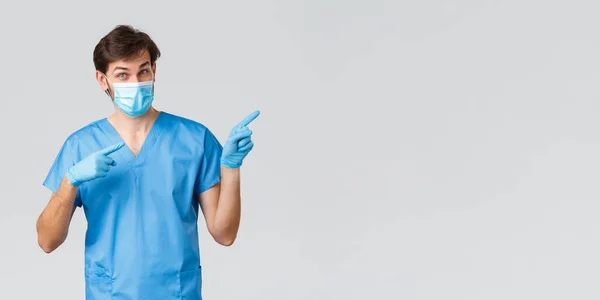 Covid-19, healthcare workers and hospital concept. Doctor inviting look at advertisement, take look. Nurse in medical mask, gloves and scrubs pointing fingers right, showing promo, grey background — Stock Photo, Image