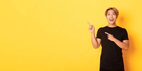 stock image Portrait of cheerful asian man pointing fingers upper left corner, smiling and showing banner satisfied, yellow background