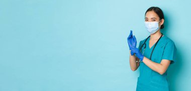Covid-19, sosyal uzaklık ve koronavirüs salgını konsepti. Gülümseyen neşeli Asyalı kadın doktor, tıbbi maske ve önlüklü stajyer, lastik eldivenler giyer, kişisel koruyucu ekipmanlar kullanırdı.