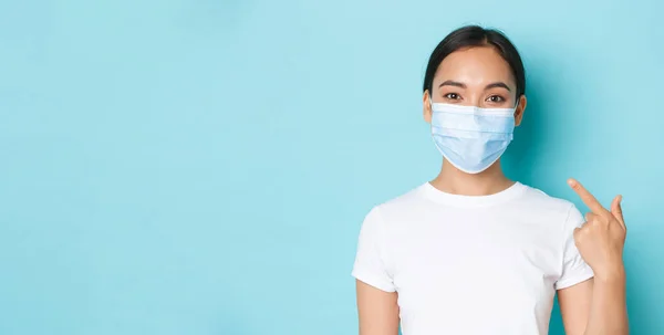 Covid-19, social distancing and coronavirus pandemic concept. Close-up of smiling asian girl in white t-shirt pointing at her medical mask on face, recommend wear personal protective equipment