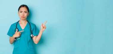 Covid-19, sosyal uzaklık ve koronavirüs salgını konsepti. Hayal kırıklığına uğramış Asyalı kadın doktor, stajyer ya da hemşire üzgün, sağ üst köşeyi işaret ediyor.